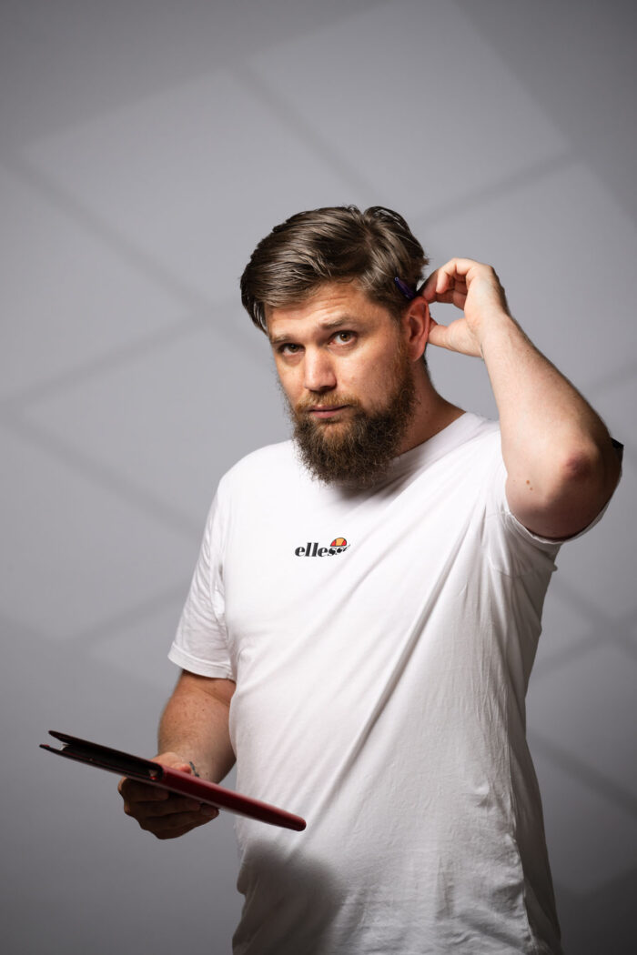 Ik sta met een notitieblok in mijn handen en kijk recht in de camera. Ik hou een pen vast die achter mijn oren steekt.